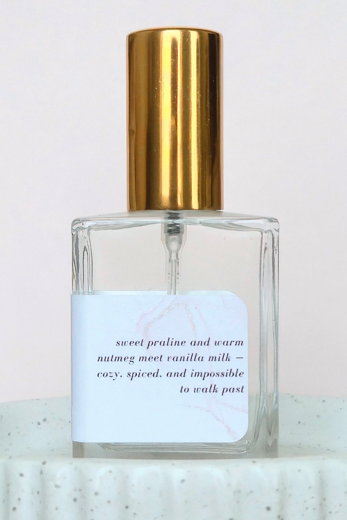 A clear glass perfume bottle with a shiny gold cylindrical cap and a white label featuring elegant black text, set against a soft light pink background.