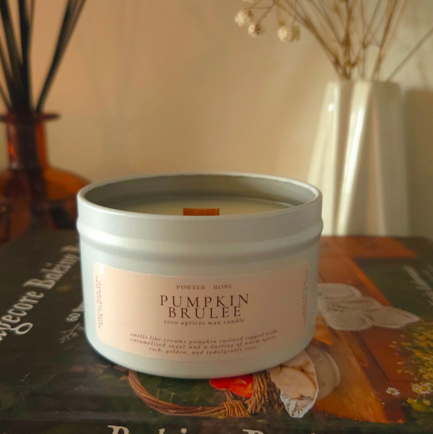 Cozy Pumpkin Brulee fall candle glowing on a decorated fireplace mantle with dried florals and cottage baking book