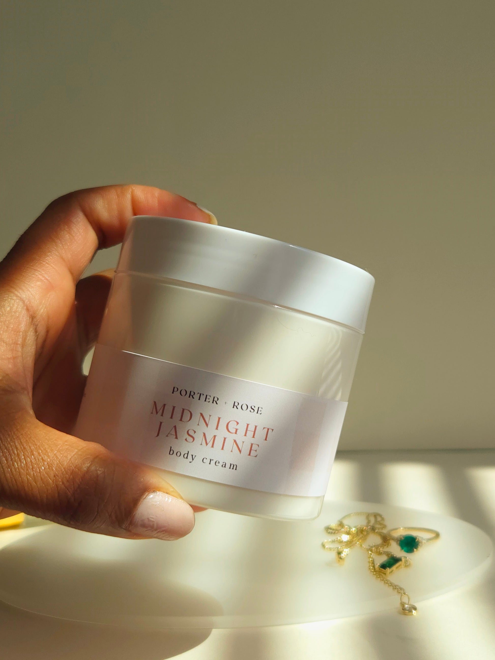 Hand holding a jar of 'Porter & Rose Midnight Jasmine' body cream with a soft background.
