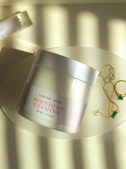Jar of Porter & Rose Midnight Jasmine body cream on a reflective surface with jewelry in the background