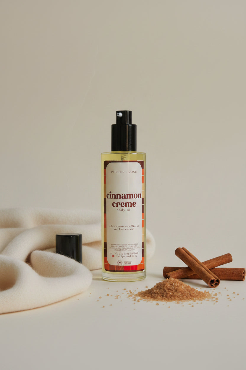 A bottle of Porter Rose Cinnamon Creme Body Oil with a black pump dispenser and a cream-colored label featuring red and orange plaid accents, set against a plain light gray background.