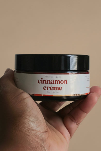 A hand holding a small jar of cinnamon creme against a plain light gray background.