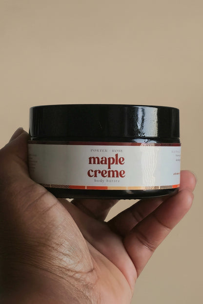A hand holding a black jar of maple creme body product against a plain light gray background.