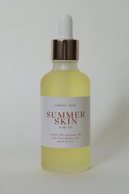 A dropper bottle of Summer Skin body oil with pale yellow gel inside, a white label, and a metallic gold collar with a white dropper cap, set against a plain light gray background.