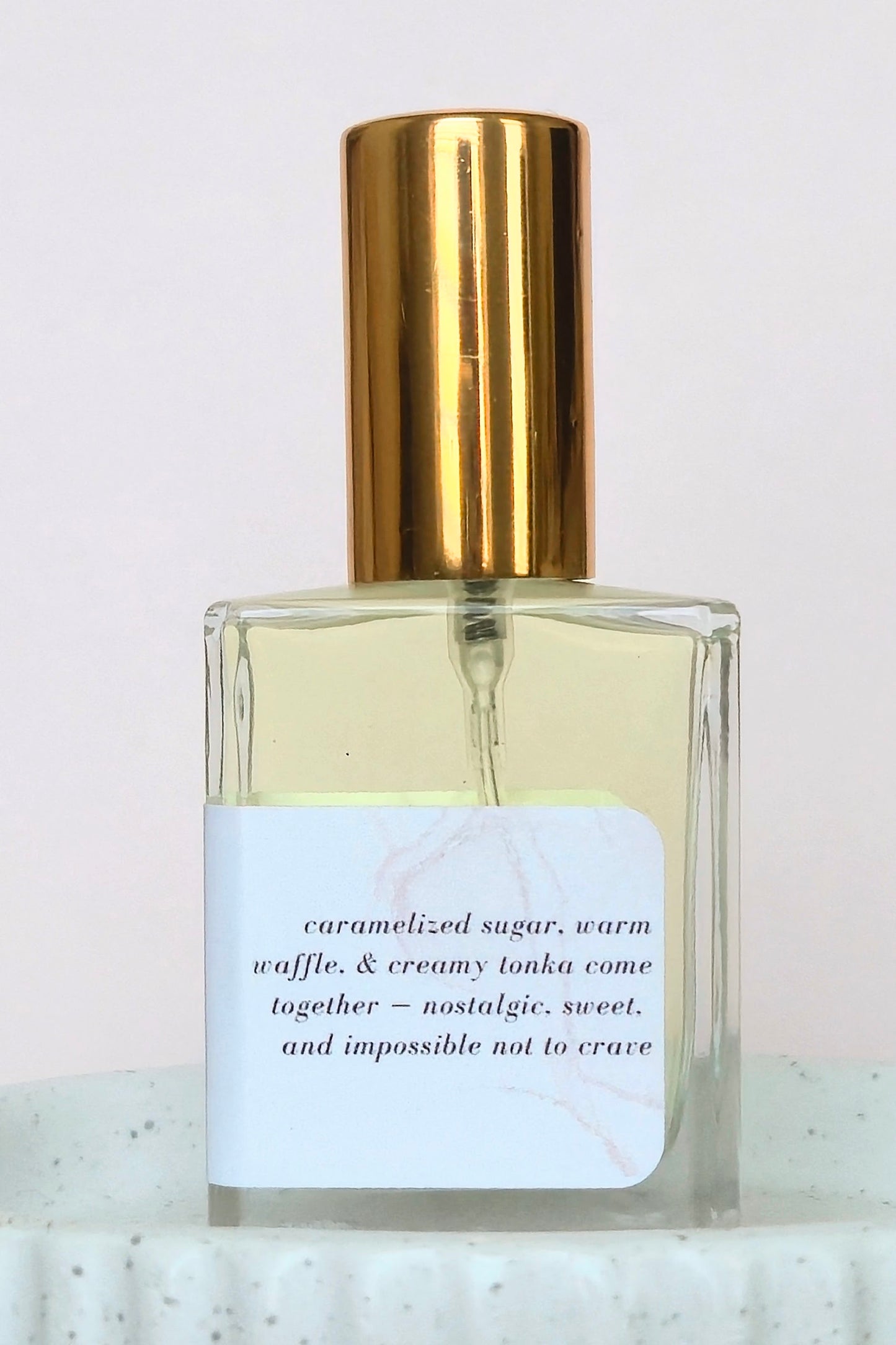 A clear glass perfume bottle with pale yellow liquid, a shiny gold cylindrical cap, and a white label with black text, set against a light background.