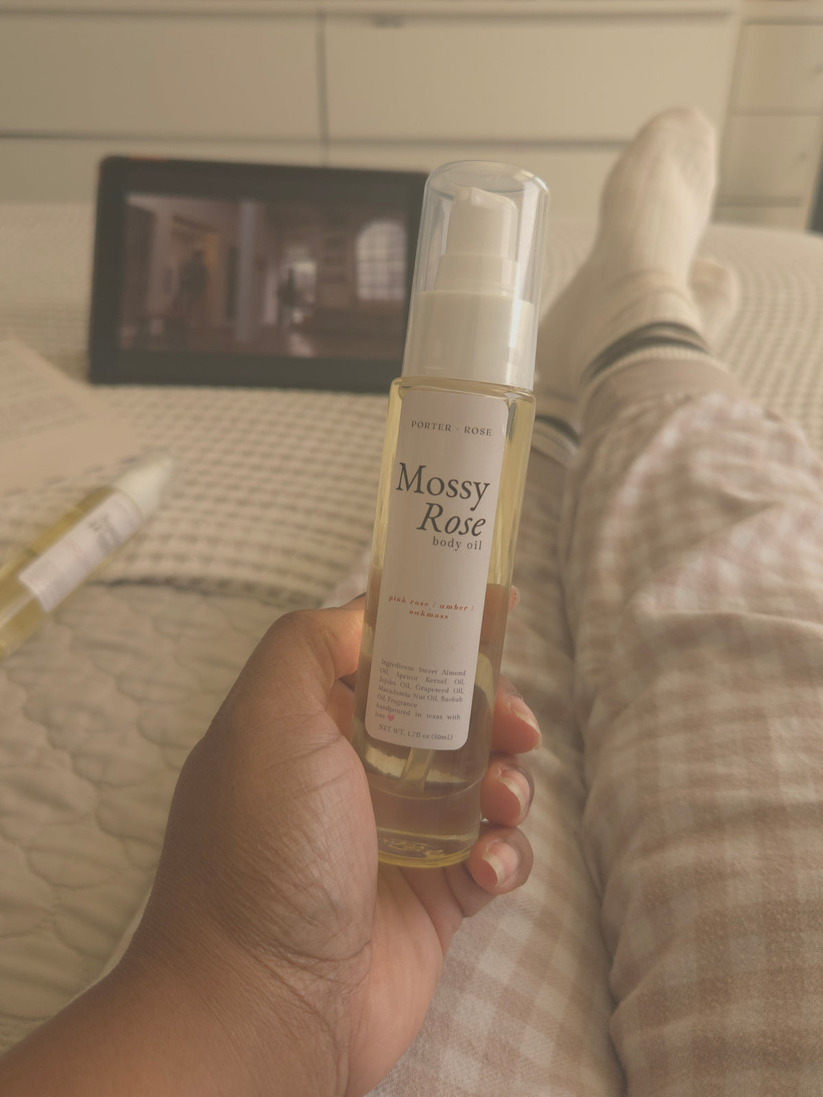 A hand holds a bottle of Mossy Rose hair oil in a bedroom setting, with the person's legs and feet visible in pajamas in the background.