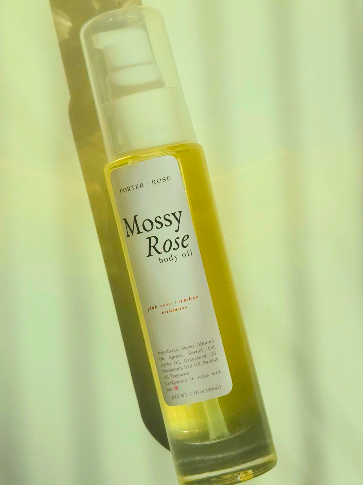 A bottle of Porter Rose Mossy Rose body oil, filled with yellow oil and featuring a white label, set against a soft, light green background.