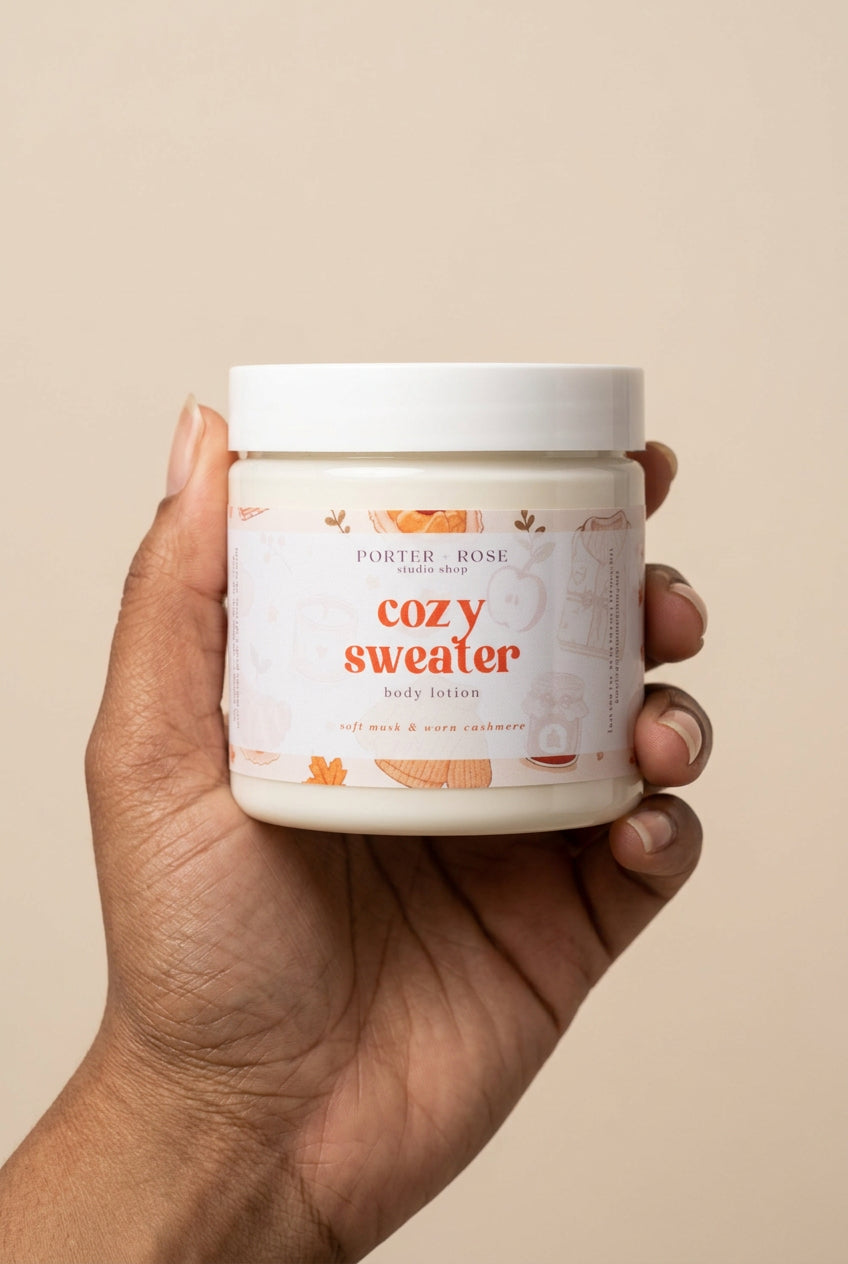 A hand holding a white jar of Porter Rose Studio Shop 'Cozy Sweater' body lotion against a plain white background.