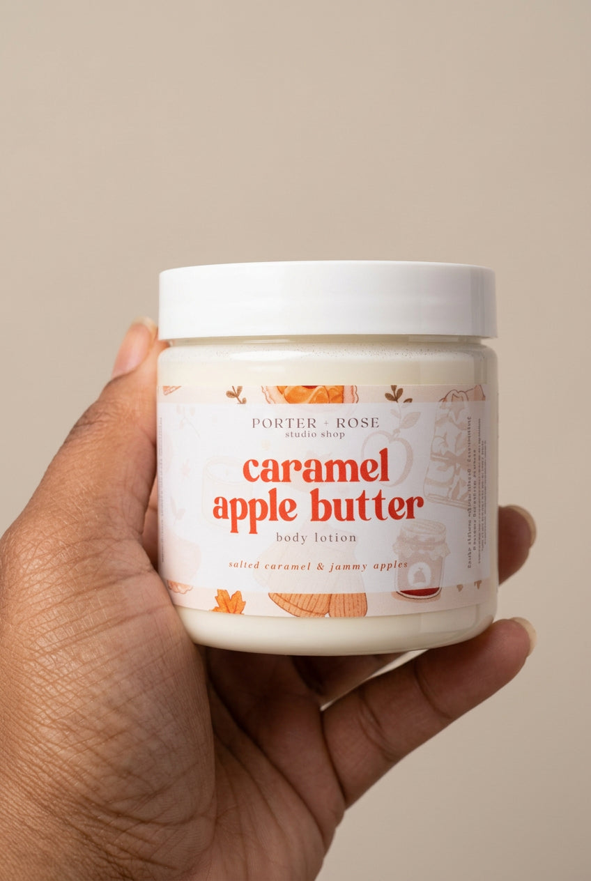 A hand holding a jar of Porter Rose Caramel Apple Butter body lotion against a bright, sunlit white background.