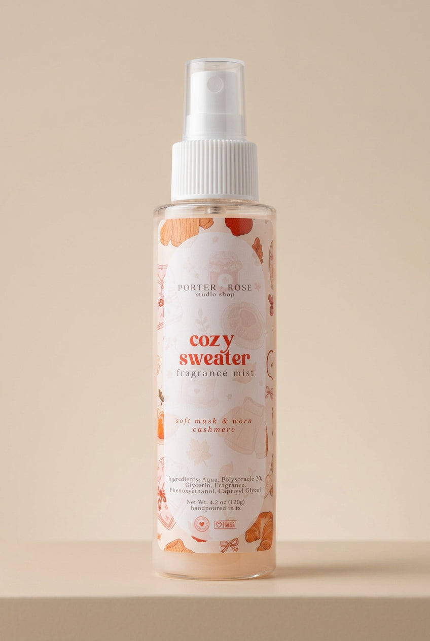 A bottle of Porter Rose 'Cozy Sweater' Fragrance Mist spray, featuring a white spray top and a clear bottle showing a light peach-colored liquid with orange floral illustrations on the label.