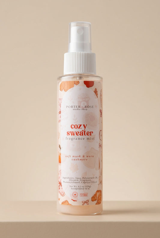 A bottle of Porter Rose 'Cozy Sweater' Fragrance Mist spray, featuring a white spray top and a clear bottle showing a light peach-colored liquid with orange floral illustrations on the label.