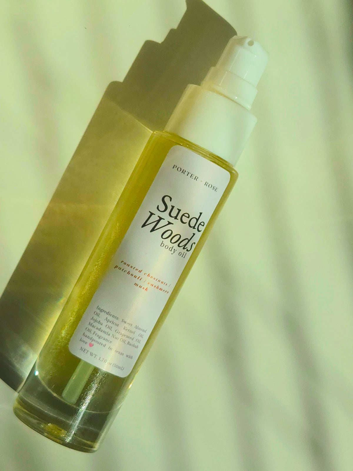 A bottle of PortedrRose Suede Woods body oil with a white pump dispenser, filled with yellow oil, set against a light background with a strong shadow.