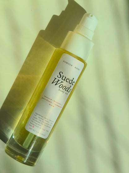 A bottle of PortedrRose Suede Woods body oil with a white pump dispenser, filled with yellow oil, set against a light background with a strong shadow.