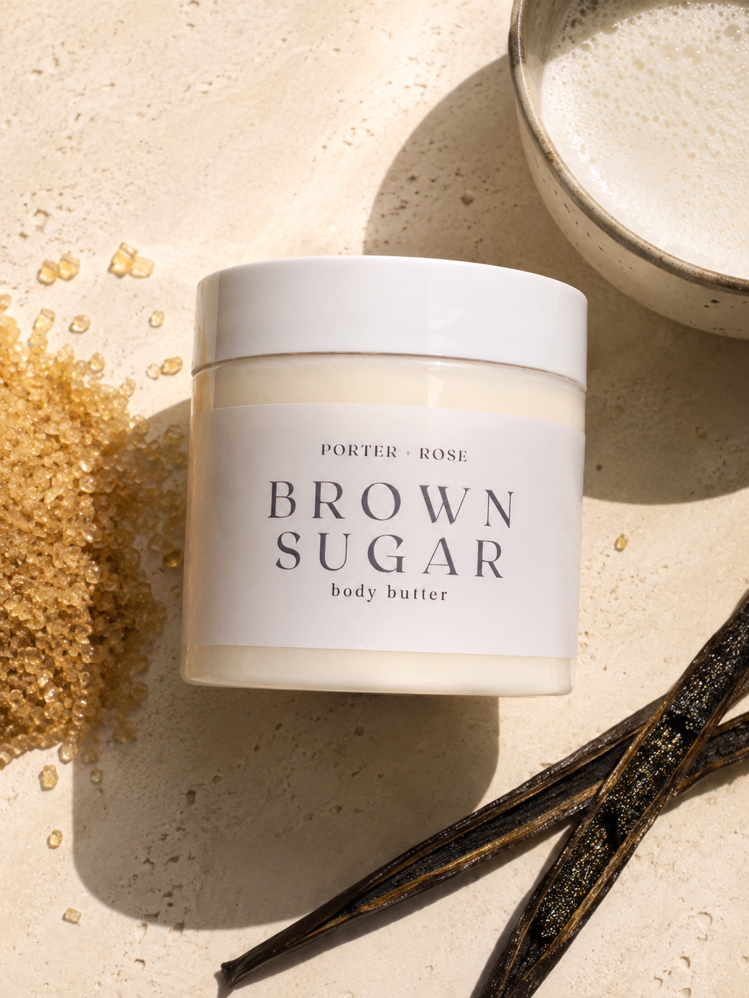 A jar of Porter Rose Brown Sugar Body Butter on a textured beige stone surface, styled with loose brown sugar, vanilla beans, and a ceramic bowl of whipped cream.