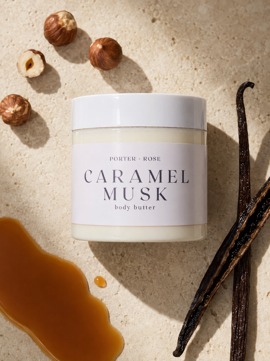 A jar of Porter Rose Caramel Musk body butter, styled on a textured beige stone surface with hazelnuts, vanilla beans, and caramel sauce.
