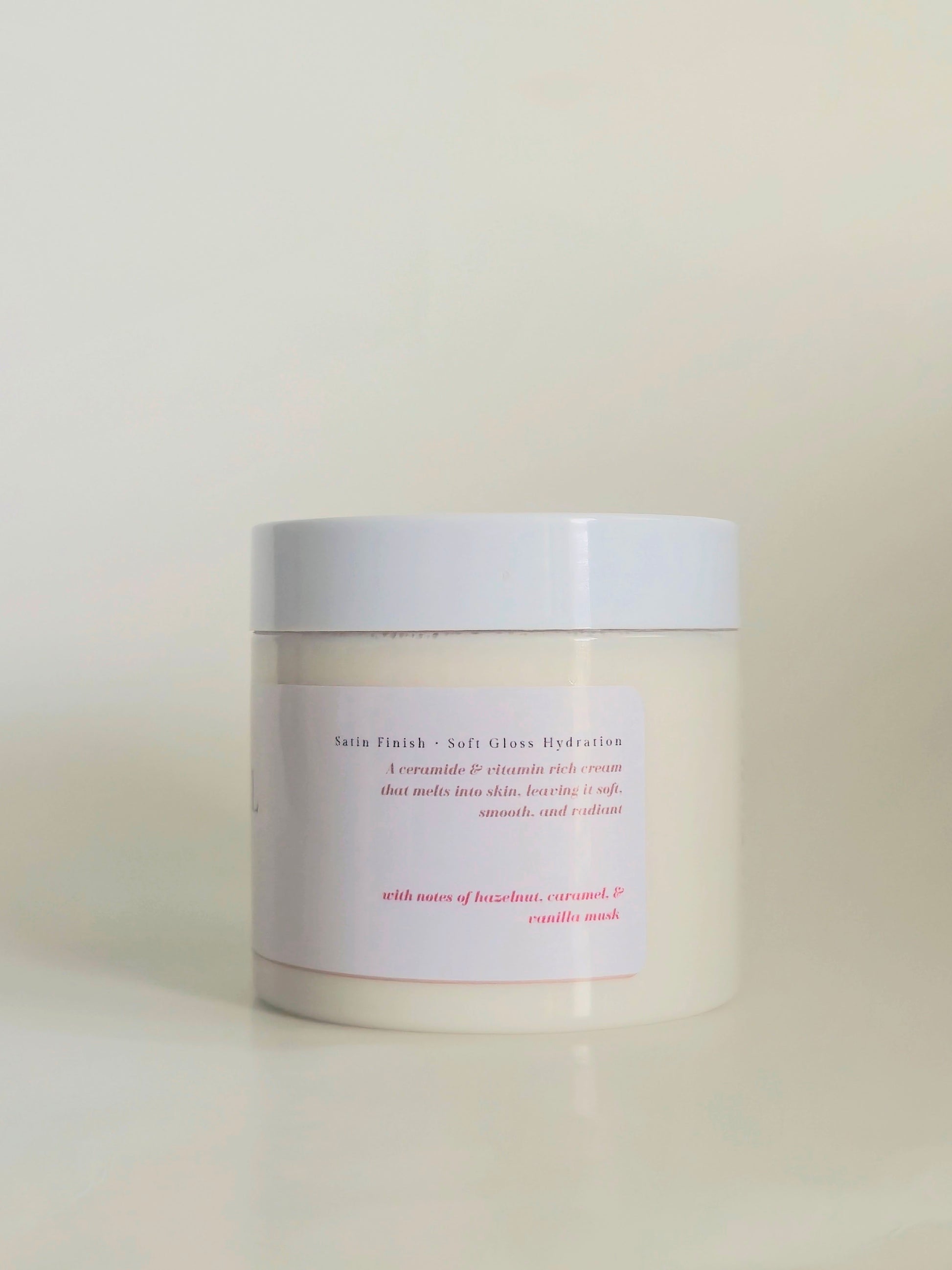 A white cosmetic jar with a white lid and a minimalist white label featuring pink text, set against a soft beige background.
