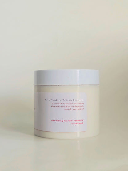 A white cosmetic jar with a white lid and a minimalist white label featuring pink text, set against a soft beige background.