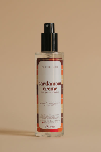 A bottle of Porter Rose Cardamom Creme Fragrance Mist with a black spray pump and a white label featuring orange and brown accents, set against a neutral light gray background.