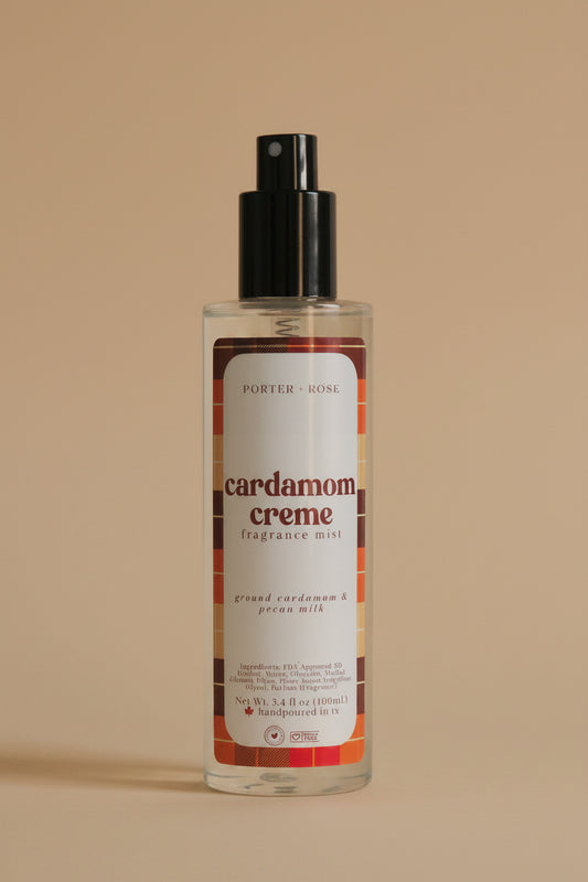 A bottle of Porter Rose Cardamom Creme Fragrance Mist with a black spray pump and a white label featuring orange and brown accents, set against a neutral light gray background.