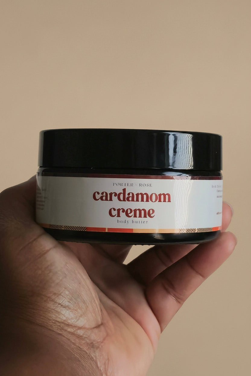A hand holding a small jar of cardamom creme against a plain light gray background.