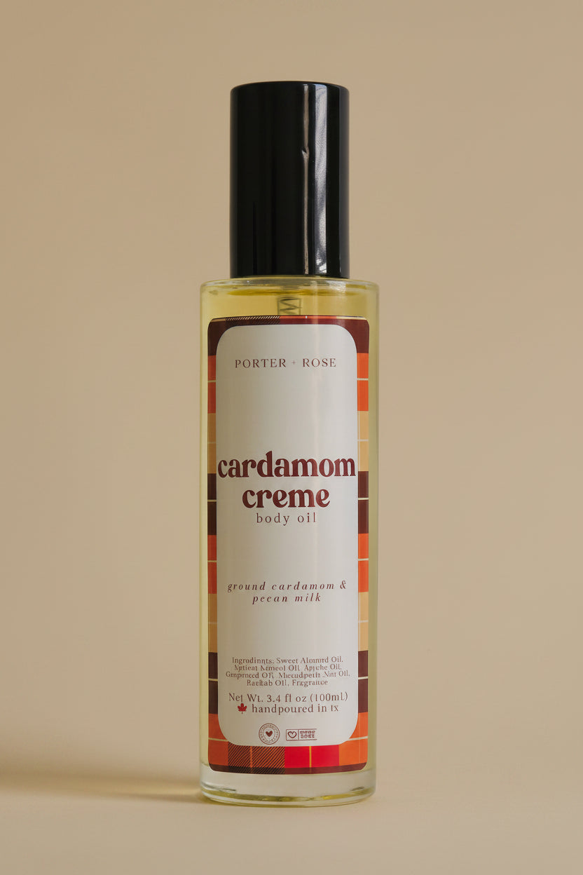 A bottle of Porter + Rose Cardamom Creme Body Oil with a black cap and a cream-colored label featuring red and brown plaid accents, set against a plain light gray background.