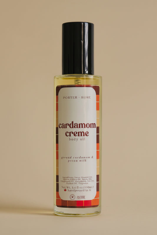 A bottle of Porter + Rose Cardamom Creme Body Oil with a black cap and a cream-colored label featuring red and brown plaid accents, set against a plain light gray background.
