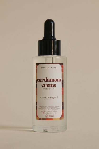 A clear bottle of Porter Rose Cardamom Creme perfume oil with a black dropper cap and a cream-colored label, set against a plain light gray background.