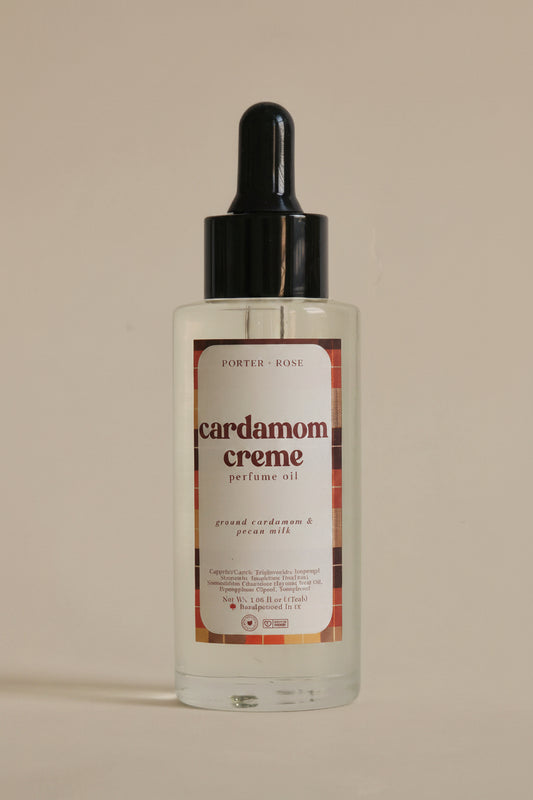 A clear bottle of Porter Rose Cardamom Creme perfume oil with a black dropper cap and a cream-colored label, set against a plain light gray background.