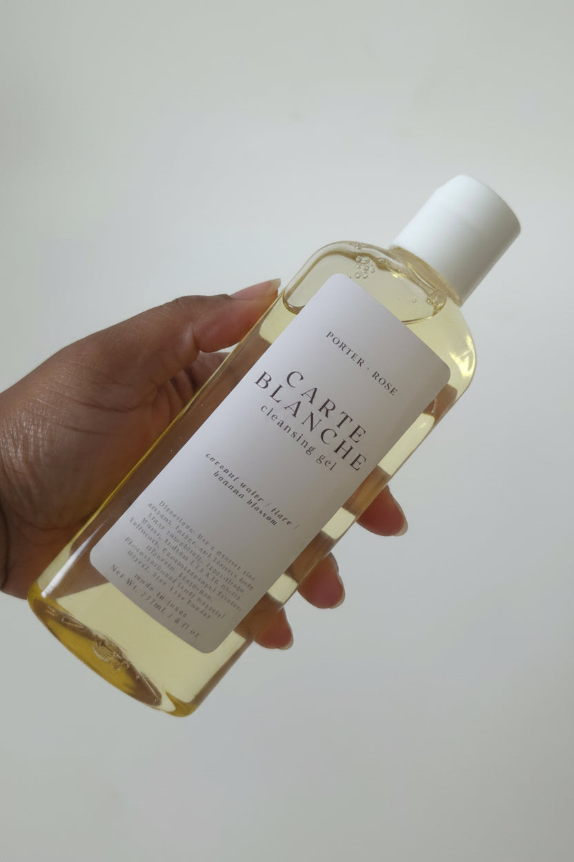 A hand holding a bottle of Porter Rive Carte Blanche Cleansing Gel, showing the pale yellow liquid inside a clear bottle with a white label and cap against a light gray background.
