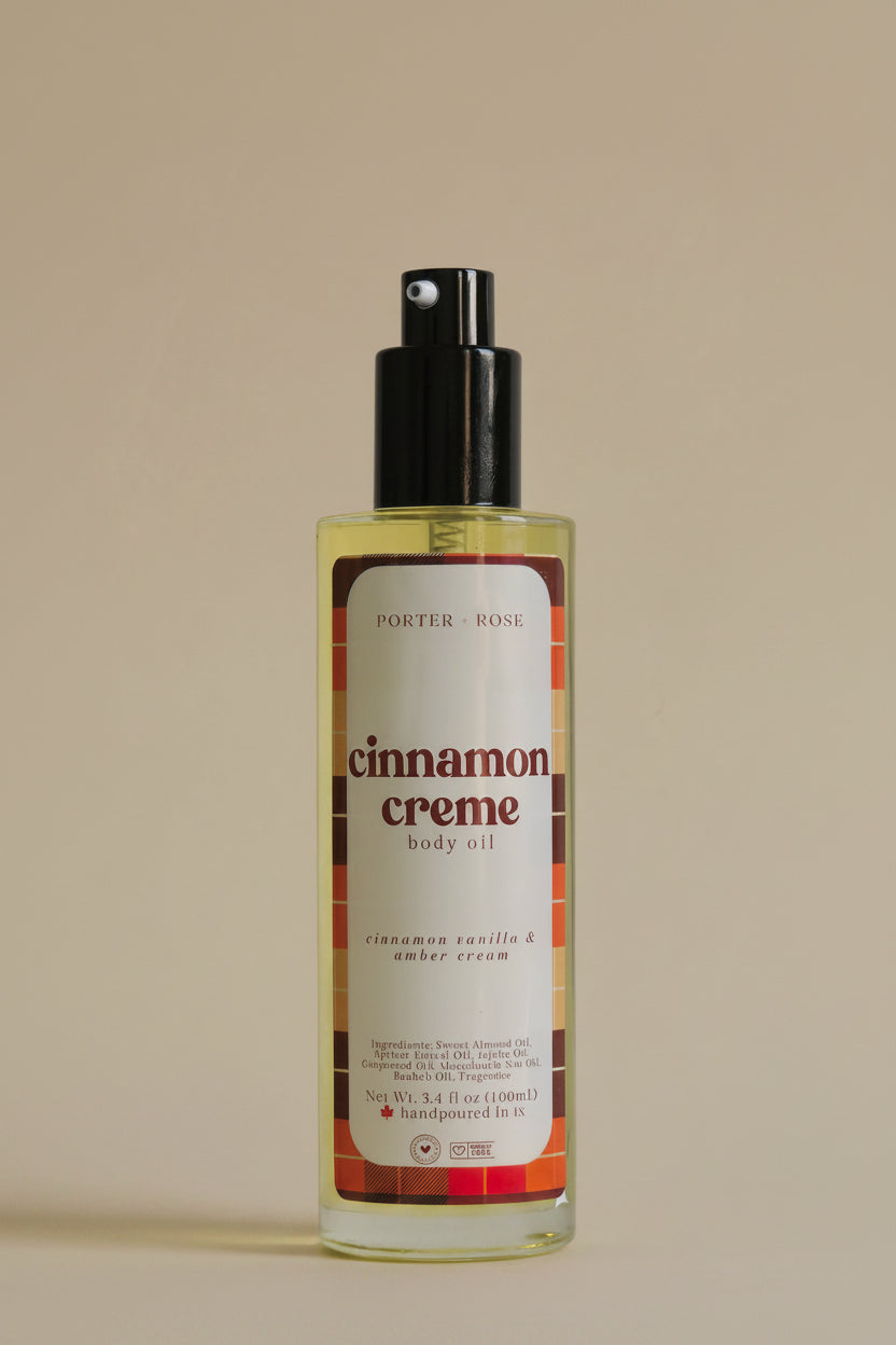 A bottle of Porter Rose Cinnamon Creme Body Oil with a black pump dispenser and a cream-colored label featuring red and orange plaid accents, set against a plain light gray background.