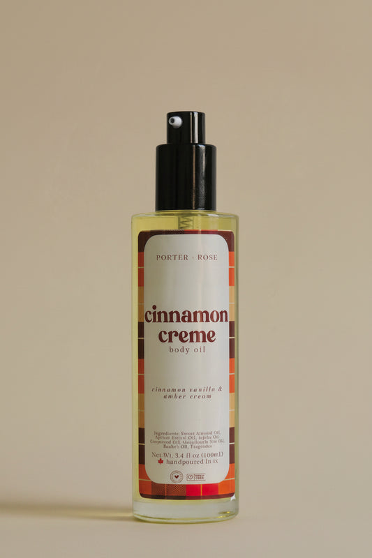 A bottle of Porter Rose Cinnamon Creme Body Oil with a black pump dispenser and a cream-colored label featuring red and orange plaid accents, set against a plain light gray background.