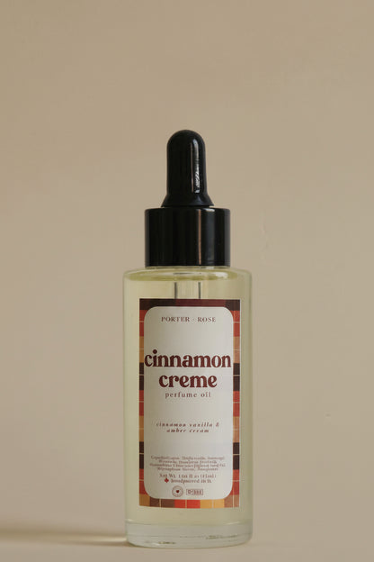 A clear glass bottle of porter rose Cinnamon Creme body oil with a black dropper cap, shown against a plain light gray background.