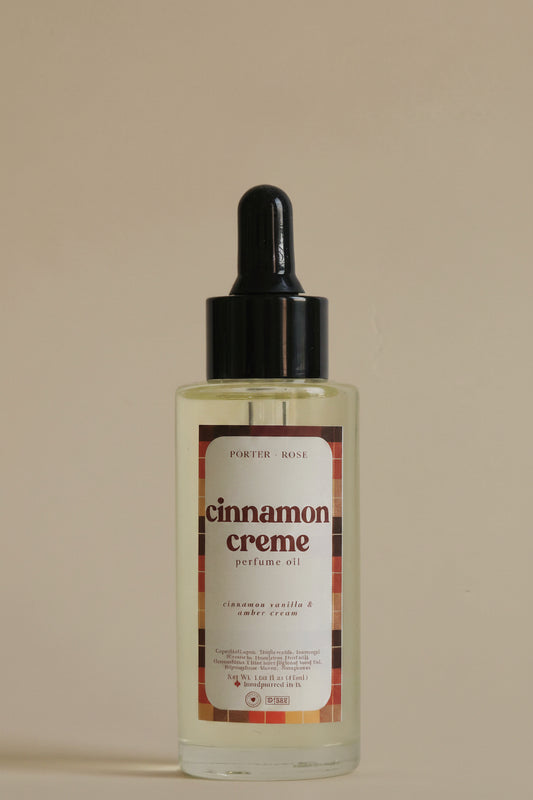 A clear glass bottle of porter rose Cinnamon Creme body oil with a black dropper cap, shown against a plain light gray background.