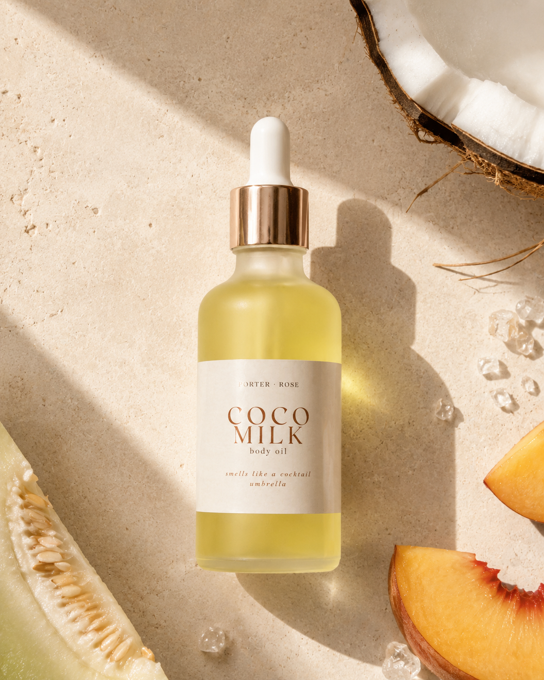 A bottle of Porter Rose Coco Milk body oil with a white dropper cap and rose gold collar, set on a textured beige stone surface with warm sunlight and shadows. Styled with halved coconut, peach slices, and ice crystals.
