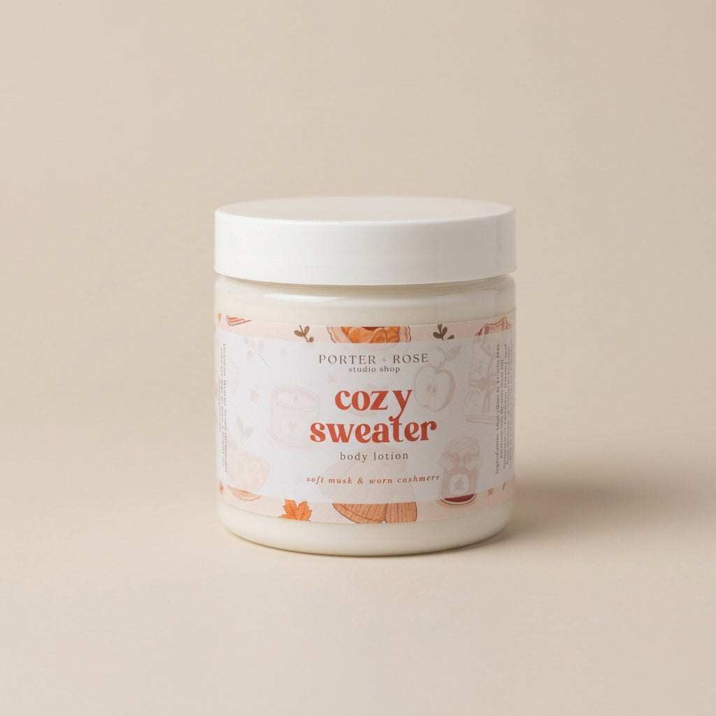 A white plastic jar of Porter Rose 'Cozy Sweater' body lotion, featuring a white lid and a label with warm orange and beige illustrations, set against a solid beige background.