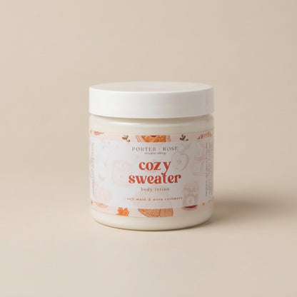 A white plastic jar of Porter Rose 'Cozy Sweater' body lotion, featuring a white lid and a label with warm orange and beige illustrations, set against a solid beige background.
