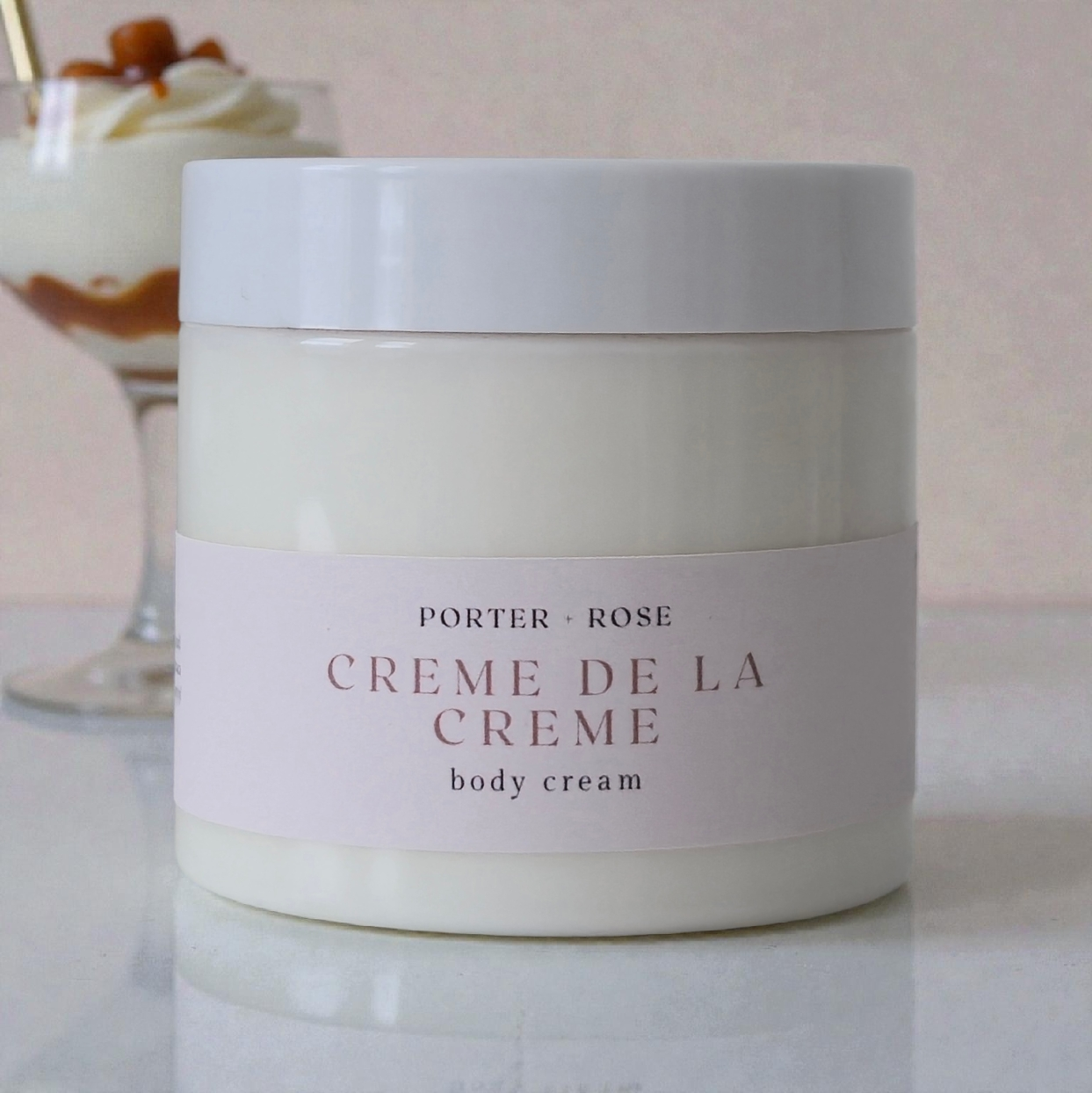 Jar of Porter & Rose Creme de la Creme body cream on a marble surface with a blurred dessert in the background.
