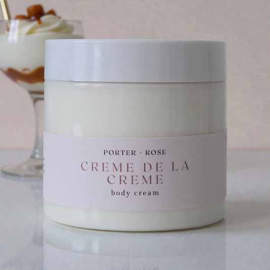 Jar of Porter & Rose Creme de la Creme body cream on a marble surface with a blurred dessert in the background.