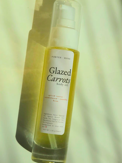 A bottle of Porter Rose Glazed Carrots body oil with a white pump and a white label, containing a yellowish-amber liquid, set against a warm, light-toned background with soft shadows.