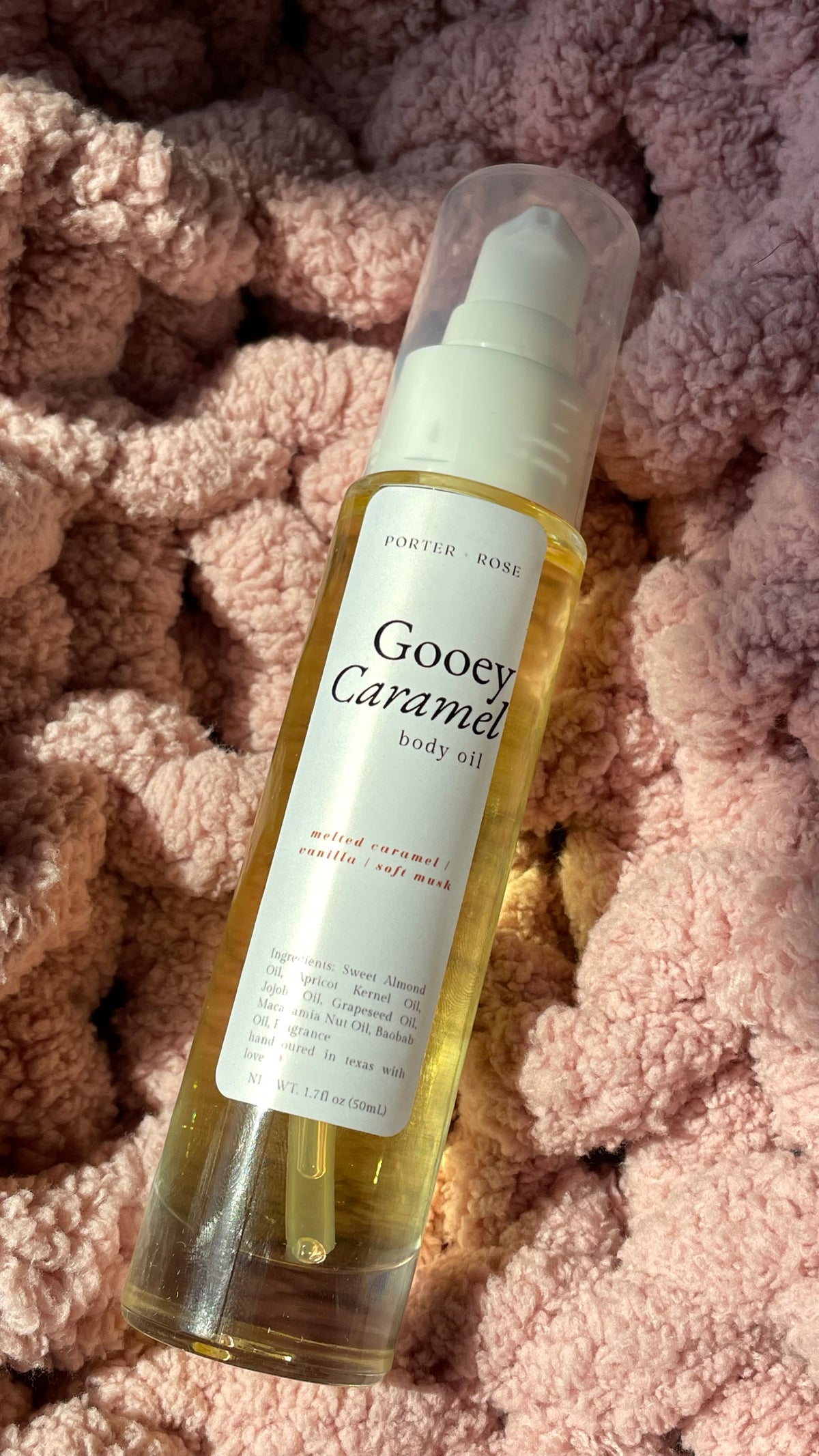 A bottle of porter Rose Gooey Caramel body oil with a clear cap and white label, resting on a textured pink blanket.