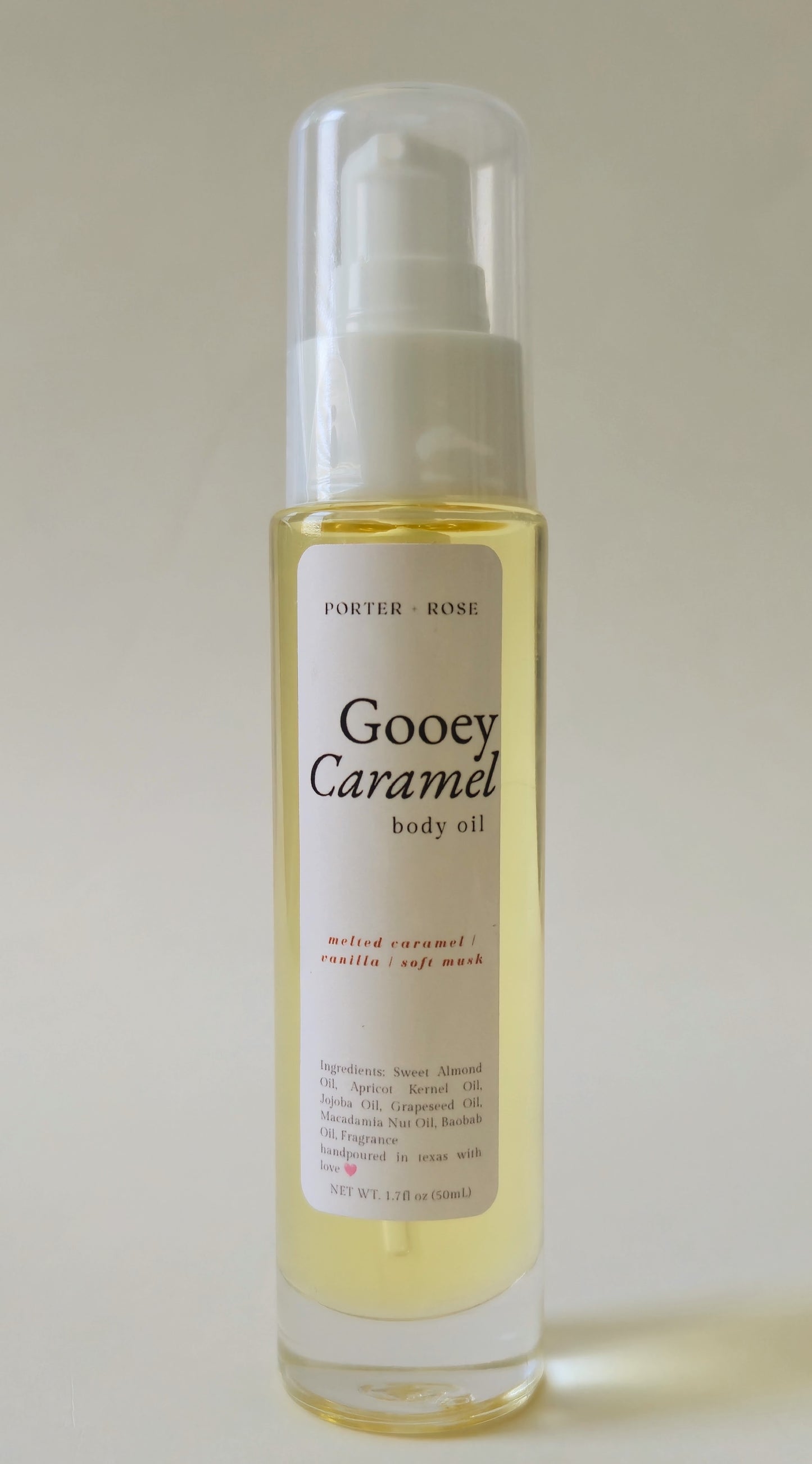 A bottle of Porter-rose Gooey Caramel body oil with a white pump dispenser and clear cap, containing a pale golden-yellow liquid, set against a plain light beige background.