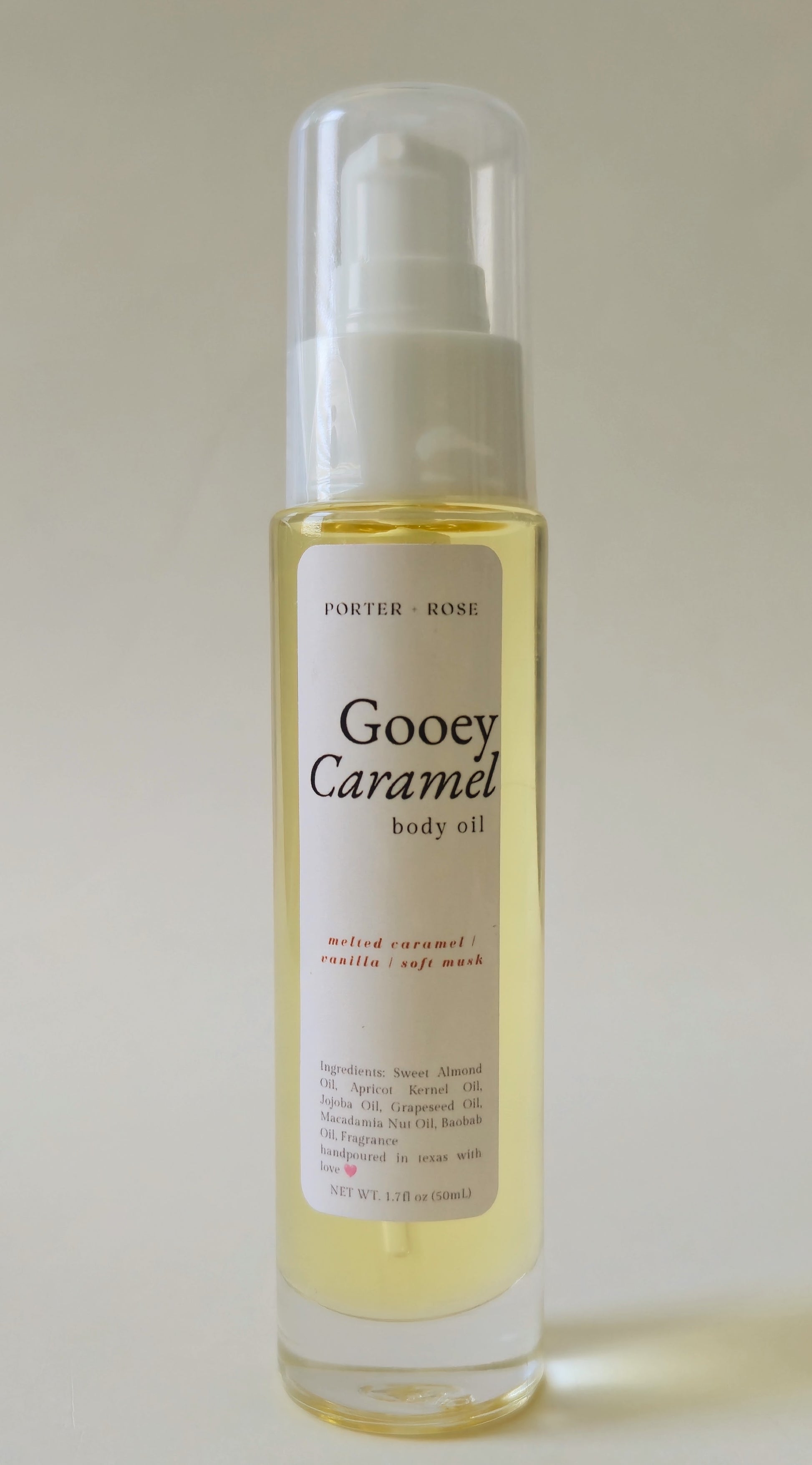 A bottle of Porter-rose Gooey Caramel body oil with a white pump dispenser and clear cap, containing a pale golden-yellow liquid, set against a plain light beige background.