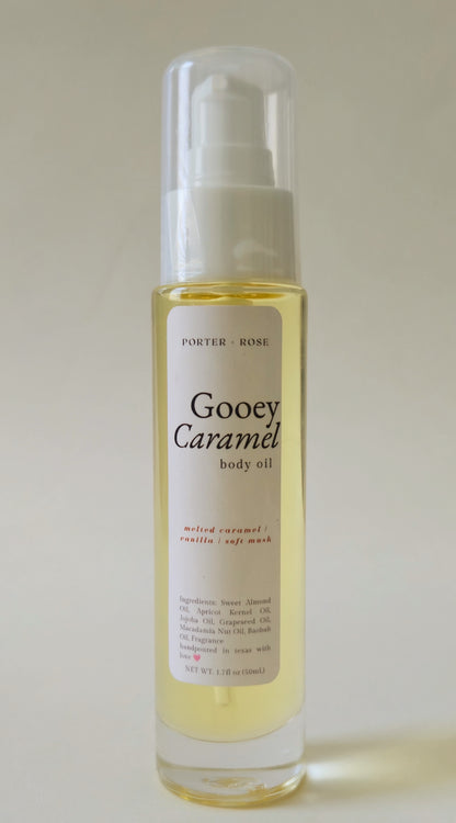 A bottle of Porter-rose Gooey Caramel body oil with a white pump dispenser and clear cap, containing a pale golden-yellow liquid, set against a plain light beige background.