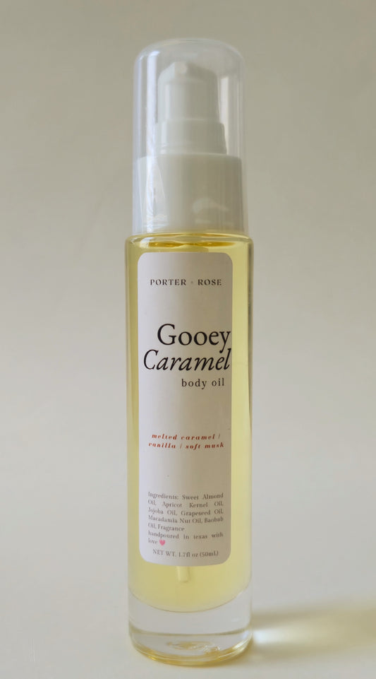 A bottle of Porter-rose Gooey Caramel body oil with a white pump dispenser and clear cap, containing a pale golden-yellow liquid, set against a plain light beige background.