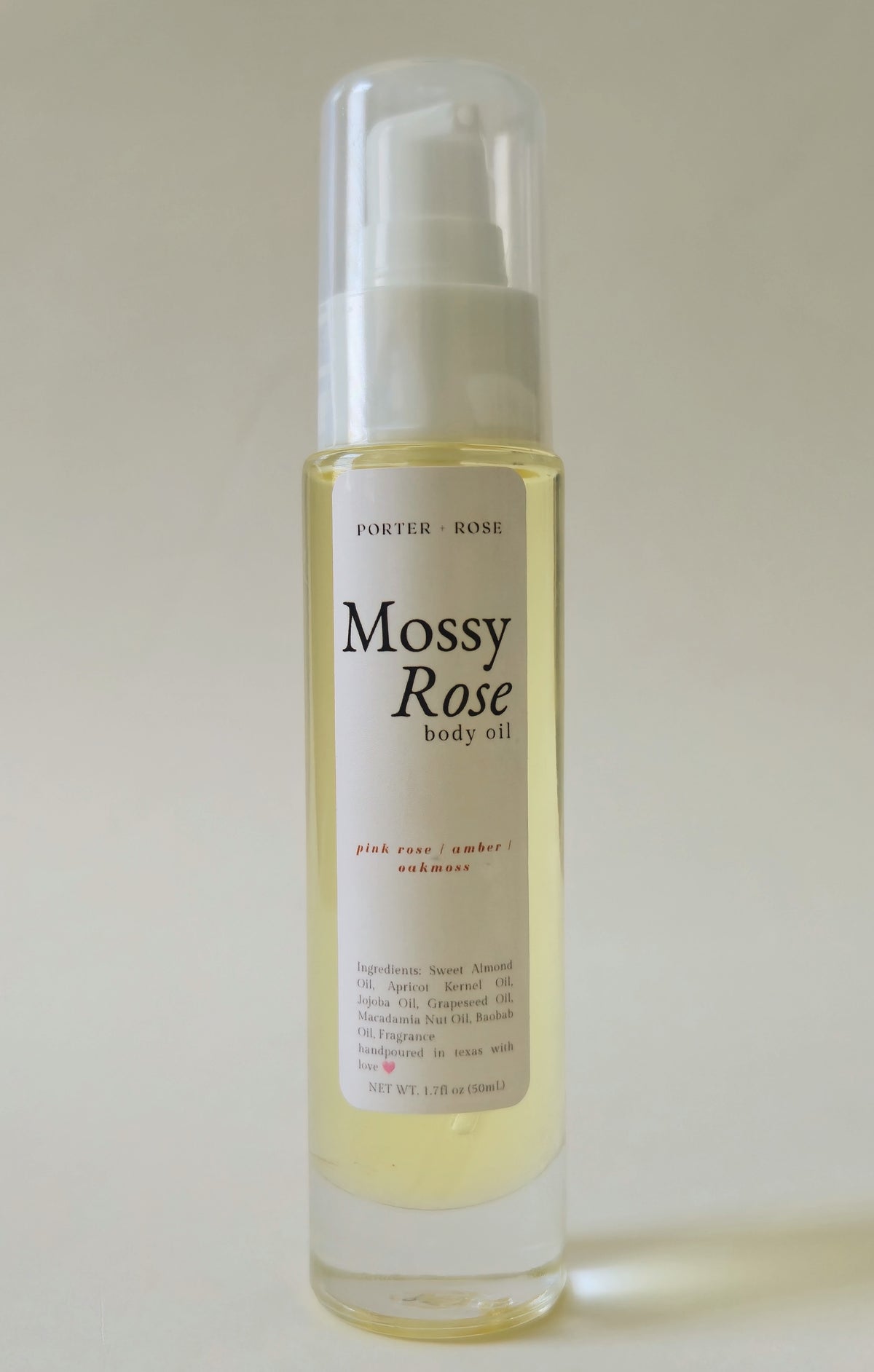 A bottle of Porter Rose Mossy Rose body oil with a white pump dispenser and clear cap, containing a pale yellow liquid, set against a plain light beige background.