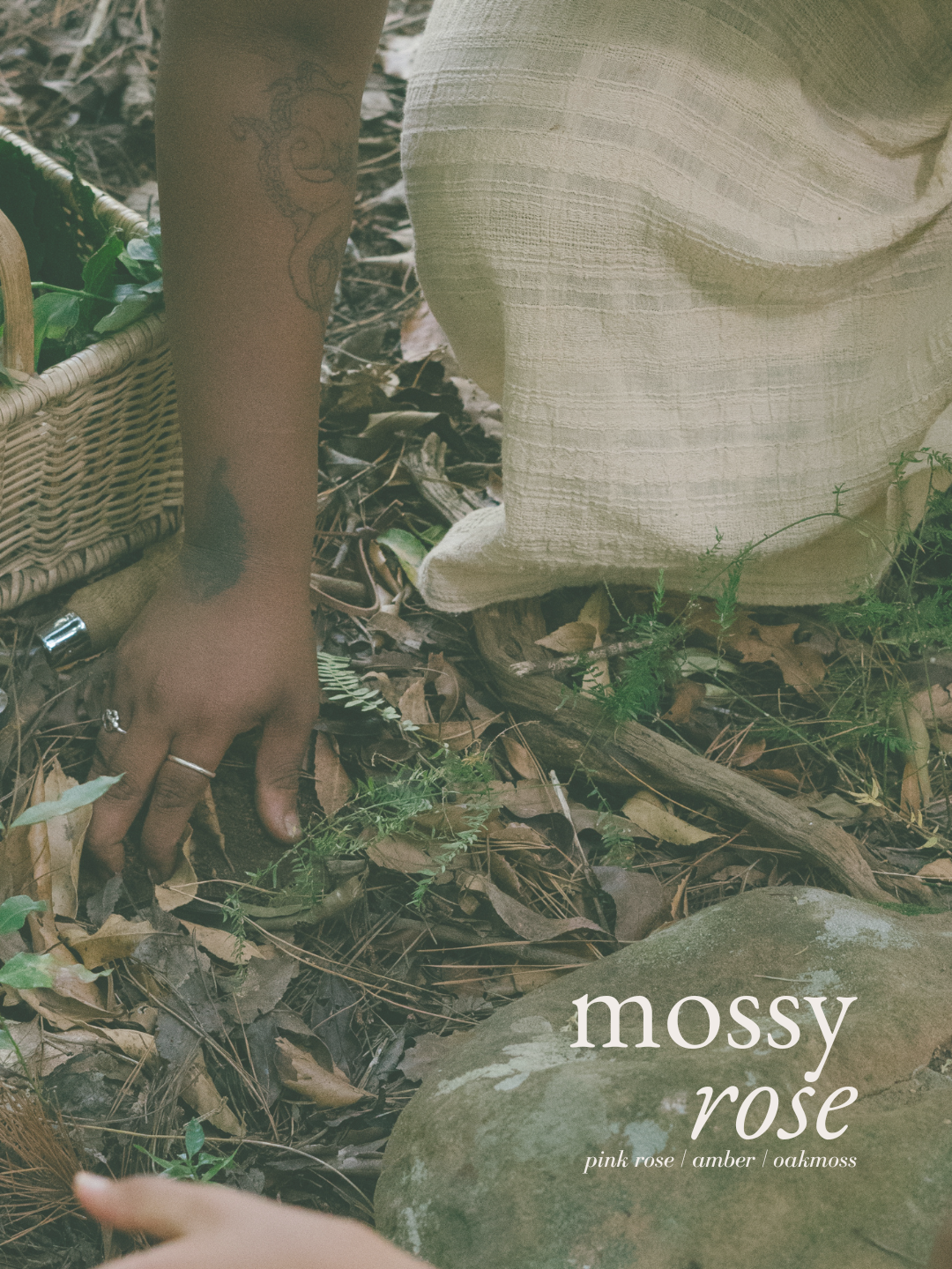 A close-up lifestyle shot of a person's hand and lower body in a rustic outdoor setting, wearing a textured beige linen garment. The scene is overlaid with the text 'mossy rose' and 'pink rose / amber / oakmoss' at the bottom right.