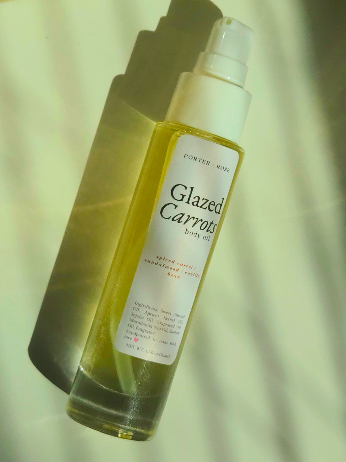 A bottle of Porter Rose Glazed Carrots body oil with a white pump and a white label, filled with yellow oil, set against a warm, shadowed cream background.