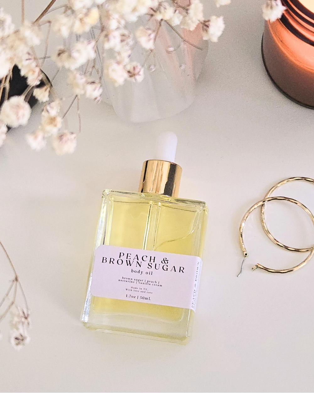 Bottle of Peach & Brown Sugar body oil with flowers and jewelry on a light surface