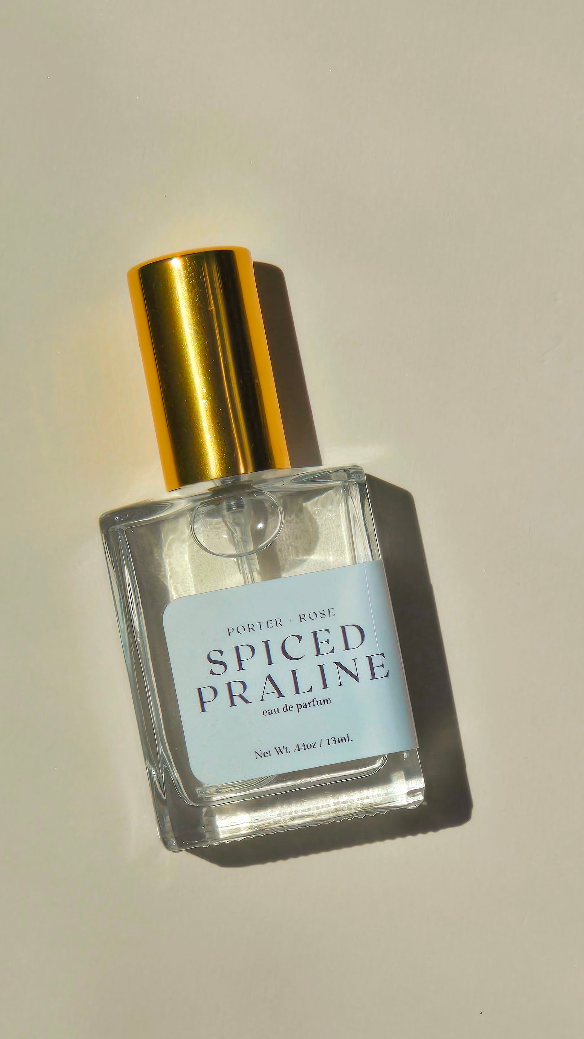 A clear glass perfume bottle with a shiny gold cap and a light blue label reading 'PORTER ROSE SPICED PRALINE eau de parfum,' photographed on a warm off-white background with a strong shadow.