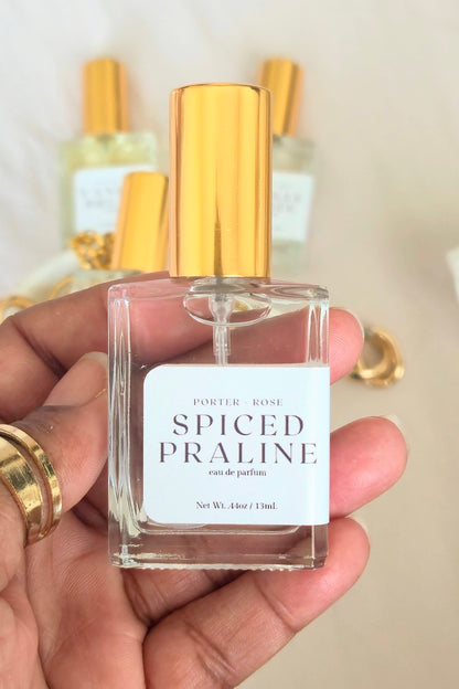 A hand holding a clear glass perfume bottle with a shiny gold cap and a white label that reads 'SPICED PRALINE' by Porter Rose. Other perfume bottles with gold caps are blurred in the background against a light neutral backdrop.